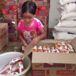 Young girl sorting food packages from a box, surrounded by stacked boxes and bags of supplies in a storage area.