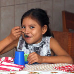 Smiling girl enjoying a meal at a table with a spoon, next to a blue cup and a checkered tablecloth.