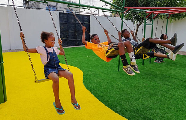 Children at the Salesian center in Ecuador playing on the swing set with smiles on their faces and the little girl is wearing a pink shirt and blue overall shorts and a boy is wearing an orange shirt